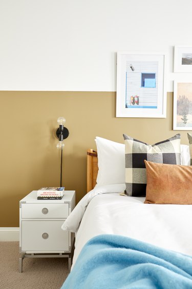 Bedroom with mustard and white color blocked wall; bed with brown and plaid pillows; white nightstand; black sconce. Art over bed.